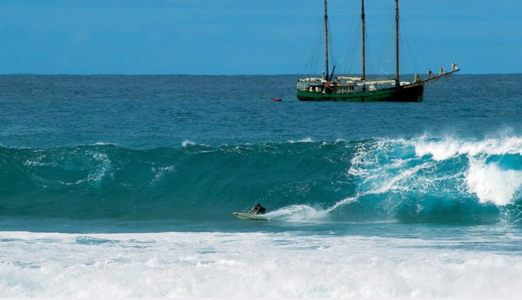 A sailboat navigating the open sea while a surfer rides a wave; ideal for illustrating surfing in Rapa Nui (Easter Island).