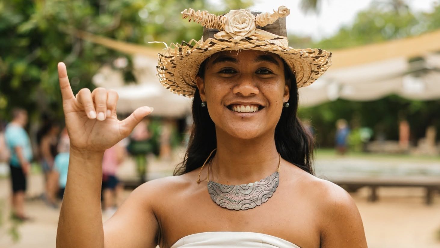 Frontal portrait of a young Polynesian woman in a white dress and woven hat throwing the shaka sign rapa nui gesture.