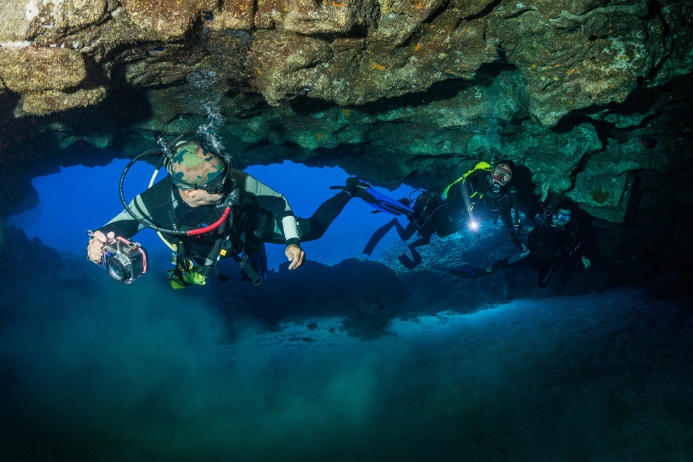 Three scuba divers, one carrying an underwater camera housing, exploring the interior of the Marau volcanic cave formation in Rapa Nui.