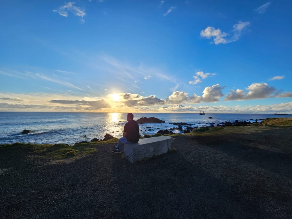 A person sitting on a stone bench facing the ocean at sunset along the rocky coast of Rapa Nui.