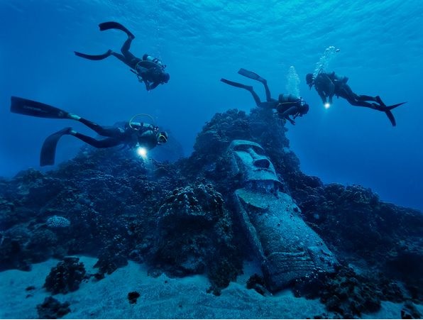 Four scuba divers with dive lights exploring a submerged moai statue leaning against a coral reef in Rapa Nui.