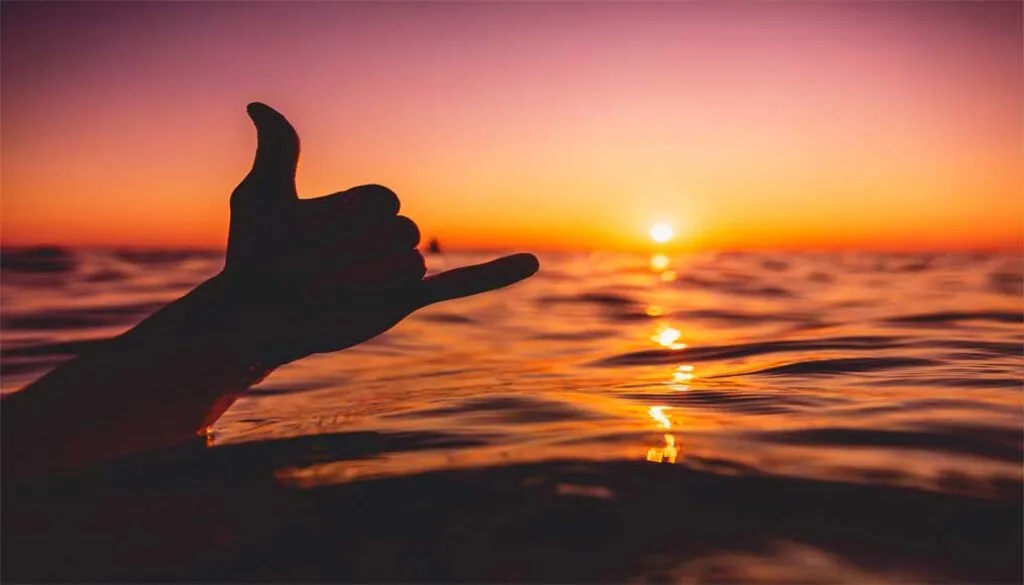 Shadow silhouette of a hand displaying the shaka sign rapa nui over the ocean during a vibrant orange sunset.