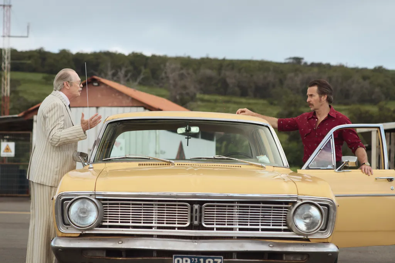 Steve Buscemi and Sam Rockwell leaning against a vintage car in an outdoor scene in Rapa Nui for Wild Horse Nine