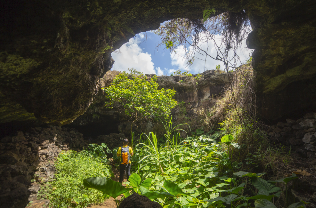 Turista bajo una claraboya natural en la cueva Ana Te Pahu rodeado de plantas.