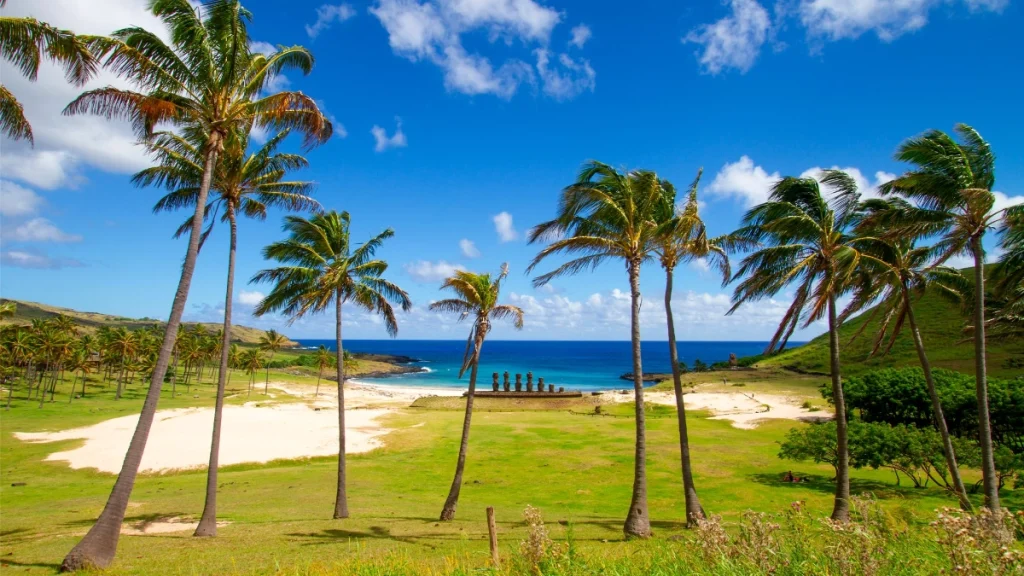 Ahu Nau Nau at Anakena viewed through palm trees, with the turquoise beach, green landscape, and blue sky in the background.