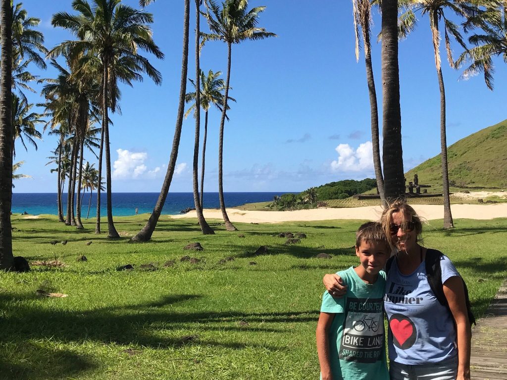 Tourists posing under tall palm trees at Anakena beach with the Pacific Ocean and a Moai platform in the background.