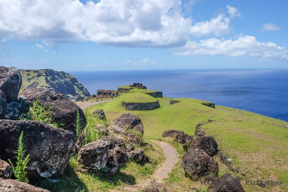 Orongo ceremonial village surrounded by green grass, with part of Rano Kau crater and the Pacific Ocean visible in the background.